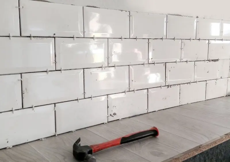 White subway tiles being laid on a wall with tile spacers and a hammer